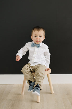 Dusty pre-tied bow tie worn by a young boy wearing white shirt, tan pants and gray shoes while sitting on a stool. 