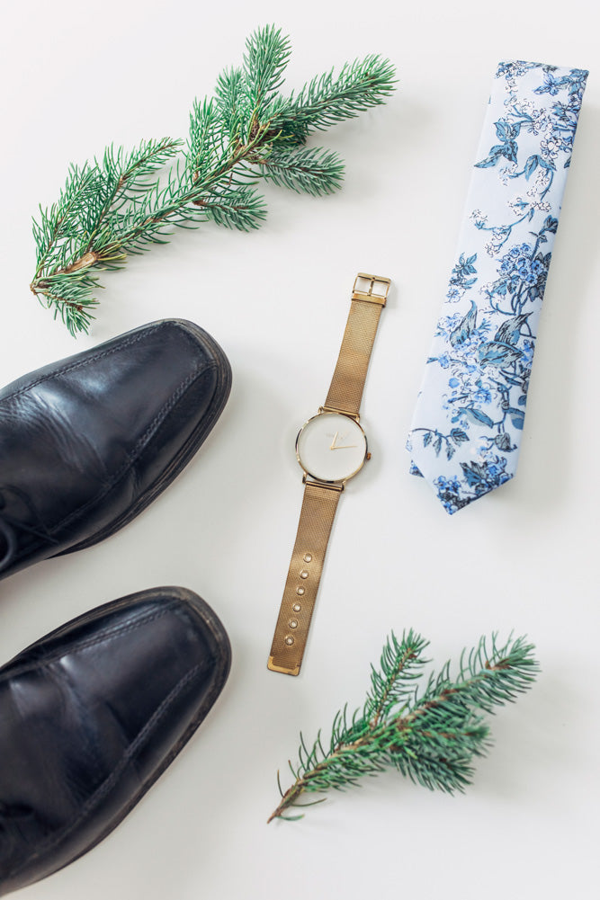 Indie Skye tie laying on the floor next to pine leaves, a gold watch and black shoes.