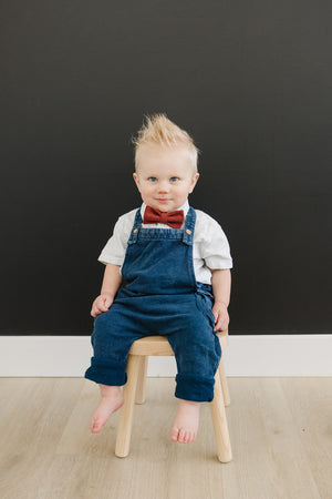Rust pre-tied bow tie worn by young boy in white shirt and blue denim overalls.