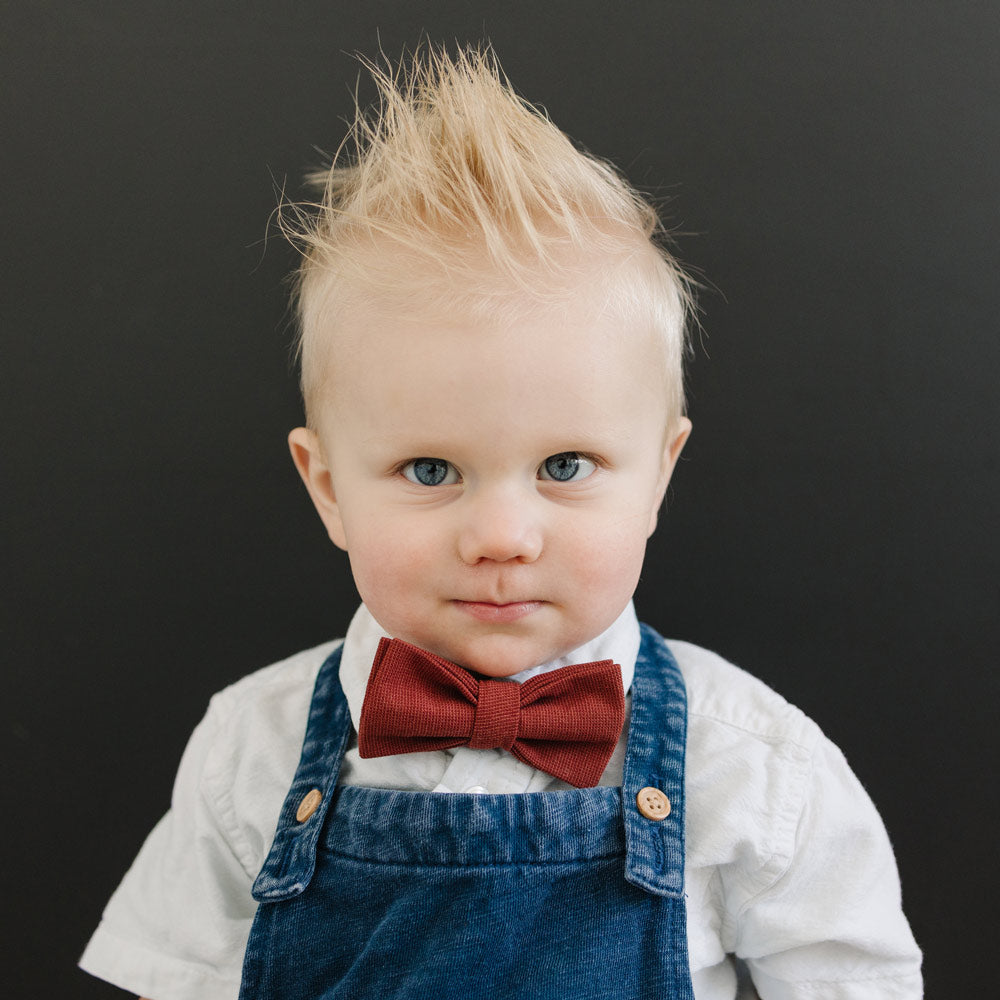 Rust pre-tied bow tie worn by young boy in white shirt and blue denim overalls.
