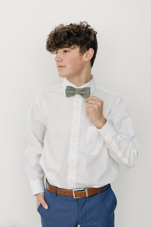 Sage pre-tied bow tie worn with a white long sleeve shirt, brown belt and blue pants. 