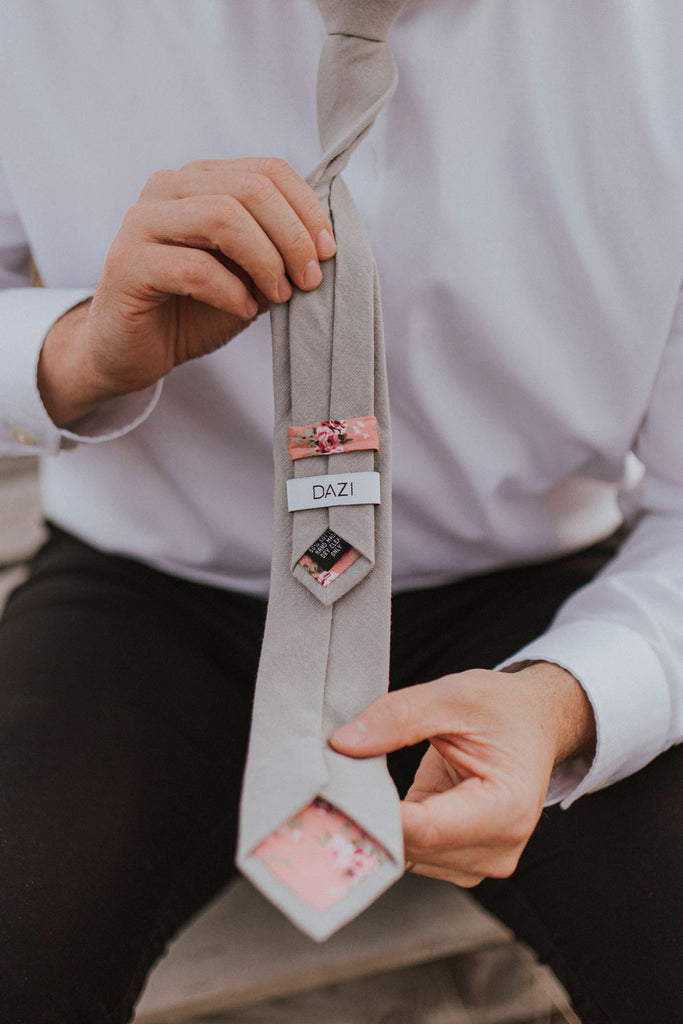 Stone tie worn with a white shirt and black pants, being held to show the back of the tie with the DAZI logo and floral lining and loop.
